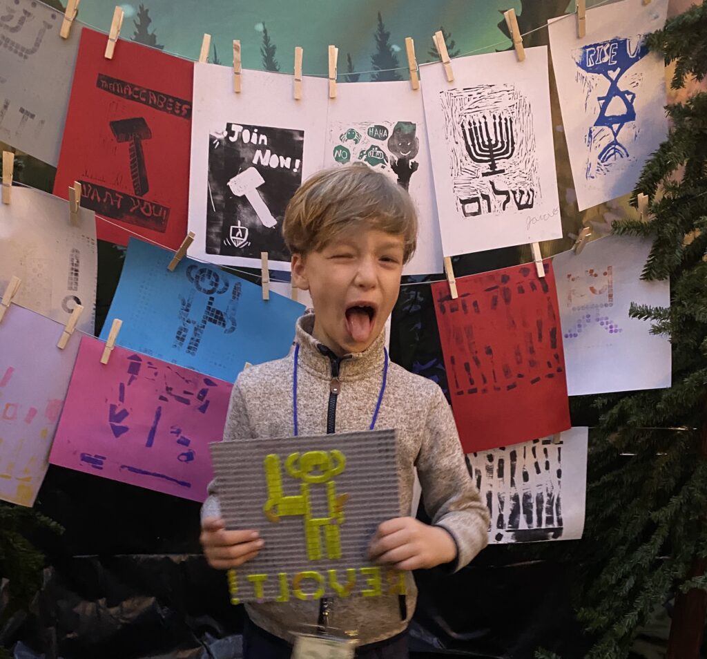 Students made Lego-dot and rubber-cut prints for Chanukah after learning about the Maccabean Revolt. This student is posing with prints and holding their Lego-dot plate at the NSP Chanukah party.